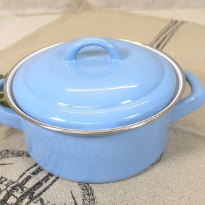 Può includere: Una pentola di smalto blu chiaro con un coperchio e due manici. La pentola ha un bordo argentato attorno alla parte superiore.