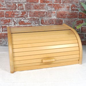 Peut inclure: Un coffre à pain en bois clair avec un couvercle roulant et une petite poignée. Le coffre est fait de lattes de bois horizontales et a des côtés arrondis. Le coffre à pain est sur une surface blanche devant un mur de briques.