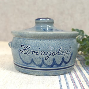 Op de afbeelding: Een blauwe keramische pot met deksel, met het woord "Heringstopf" in script. De pot heeft een getextureerd oppervlak en een golvend blauw ontwerp rond de basis. Kleine handvatten zitten aan de zijkanten.