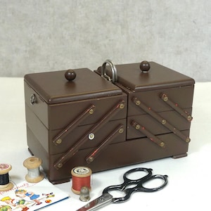 Puede incluir: Una caja de costura de madera marrón con asa, con múltiples compartimentos y tapa con bisagras. Los accesorios incluyen carretes de hilo, tijeras, un dedal y una pequeña tarjeta con ilustraciones. La caja mide aproximadamente 25 cm de ancho.