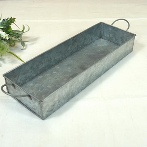 Puede incluir: Una bandeja rectangular de metal con dos asas. La bandeja está hecha de metal galvanizado y tiene un aspecto rústico y vintage.