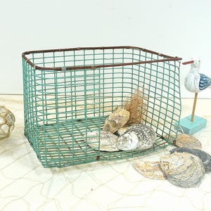 Può includere: Un cesto quadrato in filo metallico verde acqua con bordi marrone ruggine, riempito di conchiglie ed erba secca. Una statuetta di gabbiano in legno si trova nelle vicinanze su un piccolo blocco blu. Galleggianti decorativi in vetro sono visibili a sinistra.
