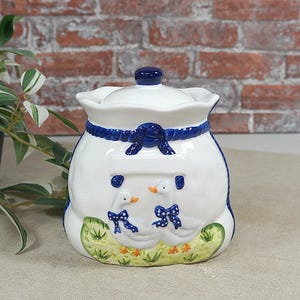Può includere: Un barattolo per biscotti in ceramica bianca con coperchio rimovibile, caratterizzato da un bordo blu a corda e un rilievo di due oche bianche con fiocchi blu a pois su una base d'erba verde con piccoli accenti di colore arancione.