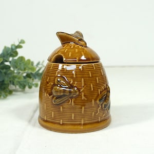 Op de afbeelding: Een bruine keramische honingpot in de vorm van een bijenkorf, compleet met een deksel en een kleine opening voor een lepel. De pot is versierd met reliëf bijen en een geweven mandpatroon. Een charmante toevoeging aan elke keuken.