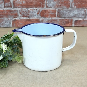 Op de afbeelding: Witte emaille kan met een blauw interieur en een donkerblauwe rand. De kan heeft een tuit en een handvat. De achtergrond omvat een bakstenen muur en groen.