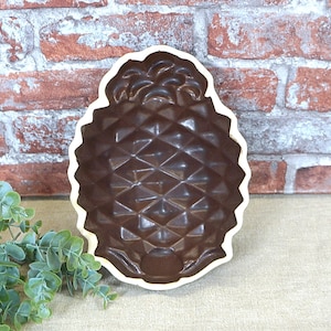 Könnte beinhalten: Eine braune, keramik-ananasförmige Kuchenform mit weißem Rand. Die Form hat eine strukturierte Oberfläche, die an eine Ananas erinnert.