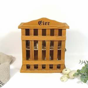 Puede incluir: Porta huevos de madera con la palabra "Eier" en la parte superior. El porta huevos es de madera clara y tiene un diseño similar a una jaula. Varios huevos moteados son visibles dentro del soporte. Una ramita de vegetación y un paño de lino están en el fondo.