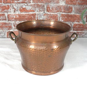 Könnte beinhalten: Ein gehammerter Kupfertopf mit zwei kleinen Griffen. Der Topf hat eine breite Öffnung und einen abgerundeten Körper. Das Kupfer hat eine warme, rötlich-braune Farbe. Der Hintergrund zeigt eine Ziegelmauer und eine weiße Oberfläche.
