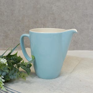 Op de afbeelding: Een lichtblauwe keramische kan met een witte binnenkant en een handvat. De kan staat op een wit linnen oppervlak.