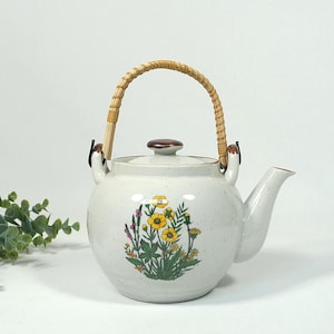 Può includere: Teiera in ceramica color crema con un design floreale con fiori gialli e viola. Ha un manico intrecciato e un coperchio marrone. La teiera è in stile vintage ed è adatta per servire il tè.