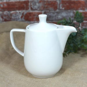 Op de afbeelding: Een witte keramische creamer met een deksel, handvat en tuit. De creamer staat op een bruin oppervlak met een bakstenen achtergrond en groen gebladerte. De creamer is ongeveer 15 cm hoog.