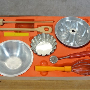 Può includere: Un set di utensili da forno giocattolo in una scatola rossa e bianca. Il set include una ciotola per mescolare, uno stampo per torta bundt, uno stampo piccolo per torte, una frusta, un tagliapasta, un cucchiaio di legno e un mattarello di legno.
