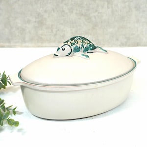 Puede incluir: Cazuela ovalada de color crema con tapa, con un asa decorativa en forma de pez. El pez está pintado en tonos verdes y blancos. El plato tiene un borde y asas verdes, adecuado para servir comida.