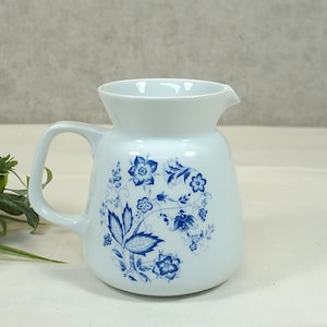 Op de afbeelding: Witte keramische kan met een blauw bloemenmotief. De kan heeft een brede opening en een handvat.