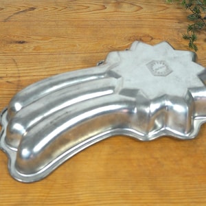 Könnte beinhalten: Eine Vintage-Backform aus silberfarbenem Metall in Form eines Kometen. Die Form hat ein sternförmiges Ende und drei gebogene Abschnitte. Die Form liegt auf einer Holzoberfläche.