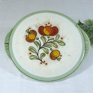 Könnte beinhalten: Eine weiße Keramik-Tortenplatte mit grünem Rand und einem Blumendesign mit orangefarbenen, roten und gelben Blüten und grünen Blättern.