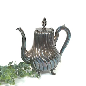 Puede incluir: Una tetera plateada con cuerpo acanalado, boquilla curva y asa decorativa. La tapa está rematada con un adorno en espiral. La tetera se apoya sobre tres patitas. El fondo es blanco.