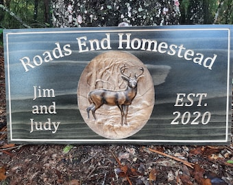 May include: A dark gray wooden sign with the text "Roads End Homestead" carved into the top. Below the text is a carved image of a deer in a forest setting. The text "Jim and Judy" is carved below the image, and the text "EST. 2020" is carved on the right side of the sign.