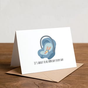 May include: A white greeting card with a blue baby car seat illustration and the text "IT'S ABOUT TO BE ARM DAY EVERY DAY." The card is on a brown envelope, set against a wooden surface.