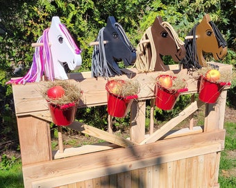 May include: A wooden horse-themed game with four horse head cutouts in white, black, brown, and tan, each with different colored manes. Red buckets with apples sit below each horse head. The structure is made of natural wood.