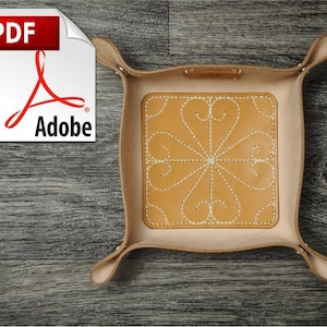 Könnte beinhalten: Ein hellbraunes Ledertablett mit quadratischer Form und vier umgeklappten Ecken. Das Tablett hat ein weiß gesticktes Design einer Blume mit sechs Blütenblättern in der Mitte. Das Tablett befindet sich auf einer Holzoberfläche.