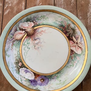 May include: A round porcelain plate featuring a pale mint green border with a gold rim. The center is white, surrounded by a decorative band of hand-painted marine life, including various seashells, a starfish, and purple sea anemones.