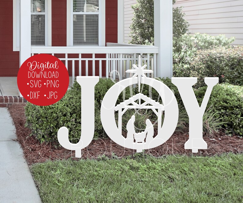 A red house with white trim. The front yard features a grass and pine straw surrounded bushes and a sidewalk. The SVG design shown is the word JOY spelled out with a nativity and star in the letter O. It is shown in white, placed in front of bushes.