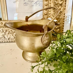 以下が含まれることがあります： 白い表面に置かれた、ハンドル付きのヴィンテージ真鍮色の金属製バケツ。 バケツは台座ベースと装飾的なディテールを備えています。 前景には緑があり、背景には額入りの書類があります。