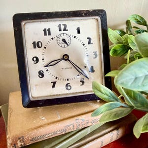 May include: Vintage square clock with a black frame and a cream-colored face. The clock features black numerals, hands, and the text "WESTCLOX". The clock is resting on a stack of old books.