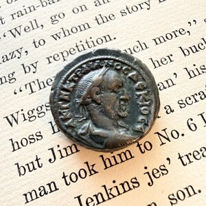 Puede incluir: Una moneda circular de metal oscuro con el perfil en relieve de una figura barbuda mirando a la derecha, rodeada de letras griegas en relieve. La moneda presenta un borde irregular y desgastado, con una superficie texturizada por el paso del tiempo.