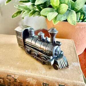 May include: A detailed, antique-style miniature train engine in shades of bronze and black. The toy train sits atop a vintage book with faded gold lettering. Green leafy plant in a terracotta pot is in the background.