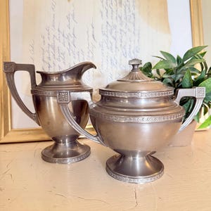 May include: A silver-toned creamer and covered sugar bowl set. The creamer has a curved spout and a handle with a decorative square element. The sugar bowl has a lid with a small knob and matching handles. Both pieces have a decorative band around the top and base.