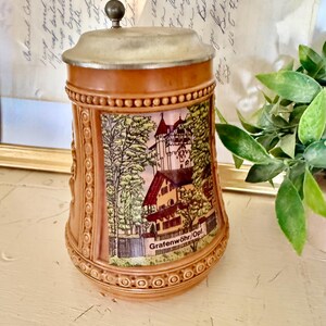 May include: A brown ceramic beer stein with a metal lid and a decorative handle. The stein features a detailed illustration of a building and the text "Grafenwöhr/Opf."