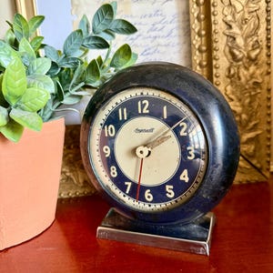 May include: A vintage Ingersoll clock with a dark blue spherical case and a cream-colored face. The clock features black numerals, a red second hand, and a silver base. A small potted plant and framed artwork are in the background.