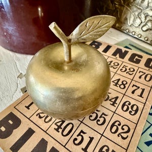 Puede incluir: Una caja de baratijas dorada en forma de manzana con un tallo y una hoja. La caja está sobre una tarjeta de bingo vintage con números negros y la palabra "BINGO". El fondo incluye un tarro rojo oscuro y una caja de metal decorativa.