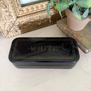 以下が含まれることがあります： 角が丸い長方形の黒い箱で、蓋には金色の文字で「WHITE」と印刷されています。蓋は、中央のテキストを囲む連続したループ状の線のパターンで構成された細い金色の縁取りで装飾されています。