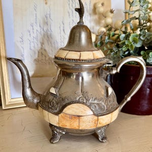 May include: A vintage silver teapot with a decorative design. The teapot features a lid with a finial, a curved spout, and a handle. The body is adorned with cream-colored rectangular inlays and floral engravings. It rests on three small feet.
