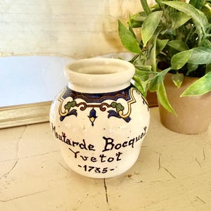 Puede incluir: Un pequeño tarro de cerámica blanquecina con una abertura ancha y una banda decorativa de diseños florales azules, amarillos y verdes. El tarro tiene una inscripción en negro que dice "Moutarde Bocq" y "Yvetot -1735-".
