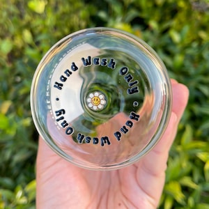 Puede incluir: Vaso transparente con las palabras "Hand Wash Only" en letras negras. Una pequeña margarita blanca con una carita sonriente amarilla está en el centro. El vaso se sostiene frente a un fondo verde borroso.