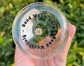 May include: Clear glass with the words "Hand Wash Only" in black lettering. A small, white daisy with a yellow smiley face is in the center. The glass is held in front of a blurred green background.