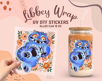 Op de afbeelding: Een glazen beker met een bamboe deksel en een helder rietje, met een blauw cartoon kat ontwerp met bloemmotieven. Een UV DTF sticker met hetzelfde ontwerp wordt naast de beker gehouden. Tekst: "Libbey Wrap".