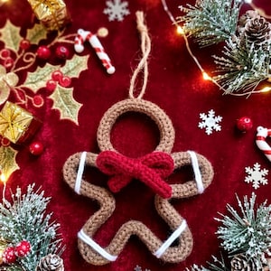 May include: A brown crocheted gingerbread person ornament featuring a red yarn bow at the neck and white yarn accents on the arms and legs. The ornament includes a jute twine loop at the top for hanging, crafted with a textured knit stitch pattern.