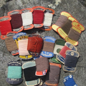 Może przedstawiać: Kolekcja starych szpulek z przędzą w różnych kolorach, w tym czerwonym, białym, brązowym i niebieskim. Szpulki ułożone są na zwietrzałej drewnianej powierzchni, z etykietami wskazującymi rodzaj i wagę przędzy w gramach.