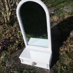 Könnte beinhalten: Weißer Bogenspiegel mit einer kleinen Schublade darunter. Der Spiegel hat einen weißen Rahmen und die Schublade hat einen runden Knopf.