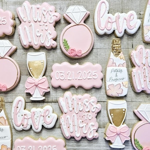 K&ouml;nnte beinhalten: Rosa und wei&szlig; glasierte Kekse mit Hochzeitsthema. Designs umfassen einen Diamantring, eine Champagnerflasche, ein Champagnerglas und die W&ouml;rter "Love" und "Miss to Mrs." Einige Kekse haben das Datum 03.21.2025.