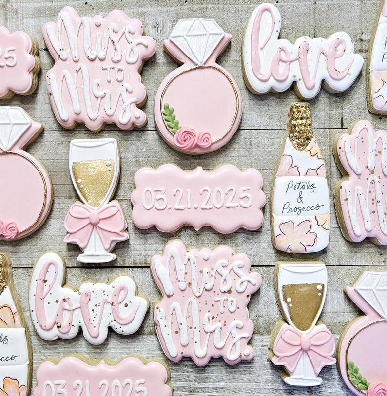 K&ouml;nnte beinhalten: Rosa und wei&szlig; glasierte Kekse mit Hochzeitsthema. Designs umfassen einen Diamantring, eine Champagnerflasche, ein Champagnerglas und die W&ouml;rter "Love" und "Miss to Mrs." Einige Kekse haben das Datum 03.21.2025.