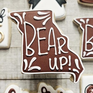 May include: A brown sugar cookie decorated with white icing in the shape of the state of Missouri. The cookie has the words "Bear Up!" written on it.