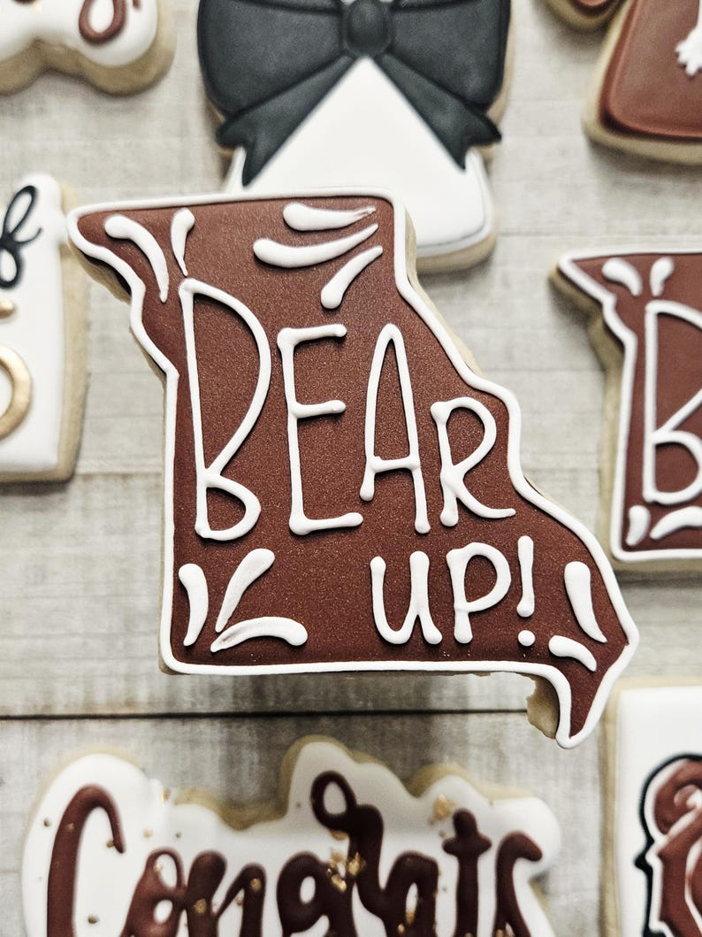 May include: A brown sugar cookie decorated with white icing in the shape of the state of Missouri. The cookie has the words "Bear Up!" written on it.