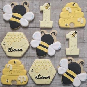 May include: A collection of decorated sugar cookies with a bee theme. The cookies include bee shapes, the number 1, and honeycomb designs. The cookies are decorated with yellow, black, white, and light blue icing. Some cookies have the name "Eliana" written on them.