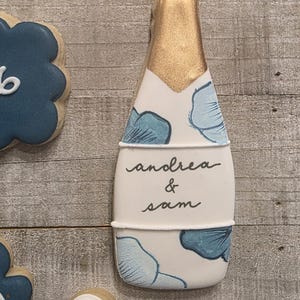 K&ouml;nnte beinhalten: Ein Keks in Champagnerflaschenform mit goldener Folienoberseite und blauem Blumendesign. Auf dem Keks stehen die Namen "andrea & sam". Weitere Kekse mit blau-wei&szlig;en Designs befinden sich im Hintergrund.